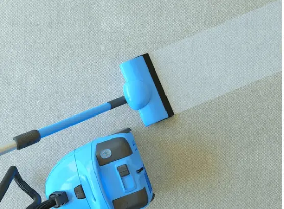 A robotic vacuum cleaner with a blue and black design is in action, cleaning a light-colored carpeted floor. The vacuum cleaner features a handle and a suction head, indicating its functionality. Its advanced design suggests it is equipped for deep cleaning, possibly with a hose attachment.